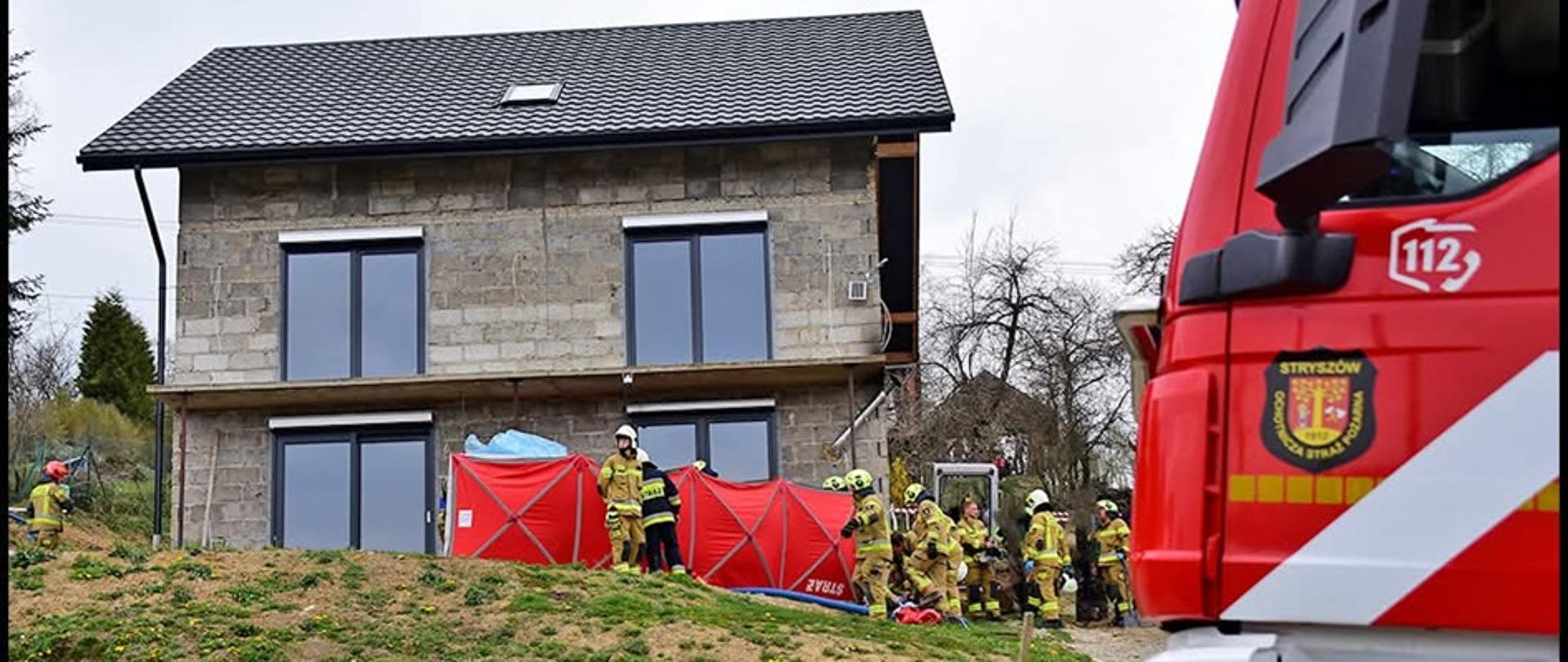Na pierwszym planie widać część kabiny samochodu pożarniczego. W tle widnieje budynek mieszkalny jednorodzinny dwukondygnacyjny. Przed budynkiem duża liczba strażaków, w sile przynajmniej dwóch zastępów, prowadzą oni działania ratownicze. Przed budynkiem znajduje się parawan z napisem Straż, zasłaniający działania strażaków, w celu ochrony prywatności osób poszkodowanych oraz odizolowania osób postronnych. 