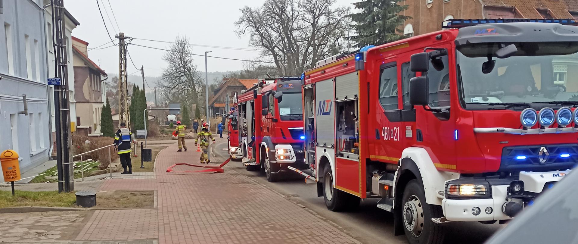 Zdjęcie przedstawia zastępy pożarnicze zaparkowane równolegle do chodnika. 