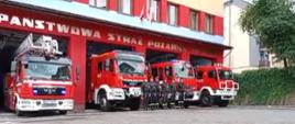 Kolorowa fotografia wykonana w pochmurny dzień na zewnątrz. Przedstawia strażaków stojących w szeregu z dowódcą oddającym honor na tle samochodów pożarniczych z włączonymi światłami dźwiękowymi i świetlnymi.
