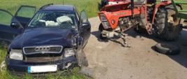 Uszkodzony samochód osobowy marki Audi oraz ciągnik rolniczy Ursus po zderzeniu czołowym. W aucie osobowym uszkodzone nadkole od strony kierowcy, lampa, zderzak oraz przednia szyba. W ciągniku urwane przednie koło. W tle samochód ratowniczo-gaśniczy.