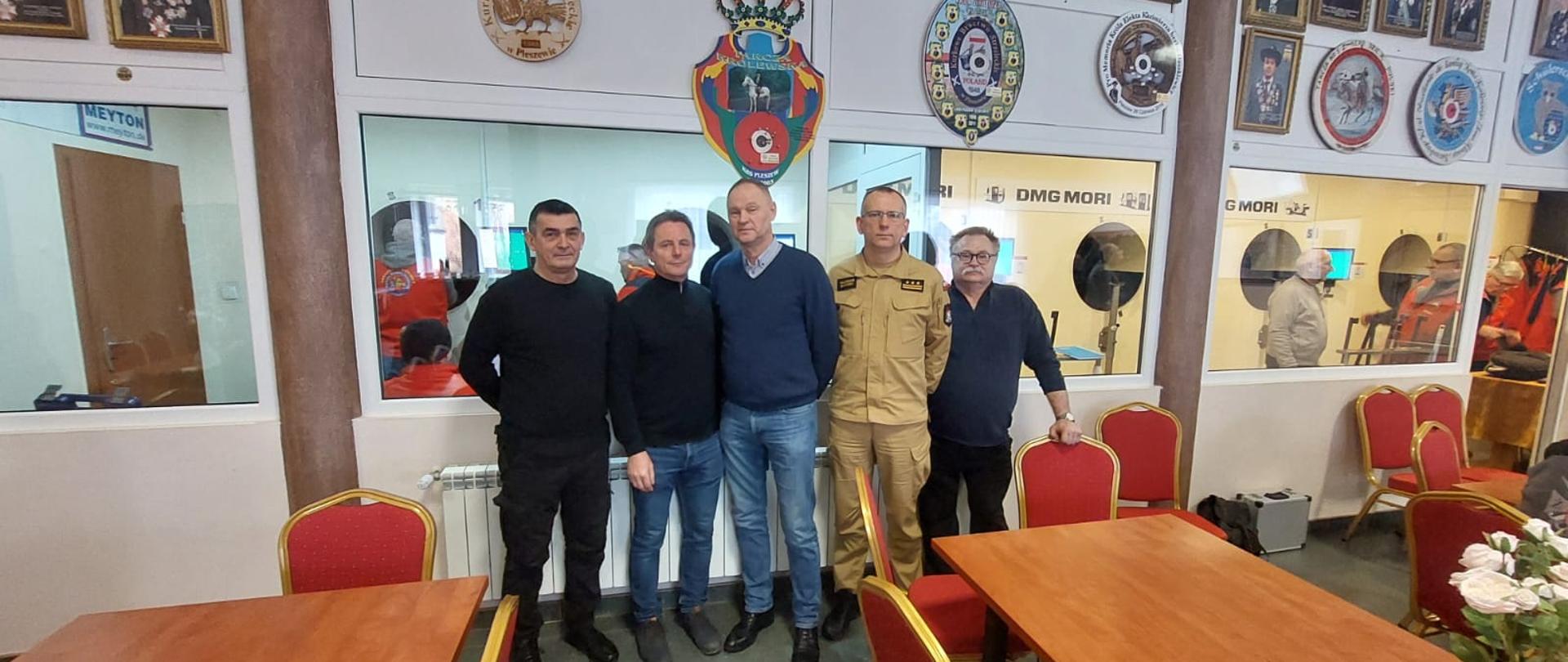 osoby stojące przy stołach a w tle strzelnica w pomieszczeniu 