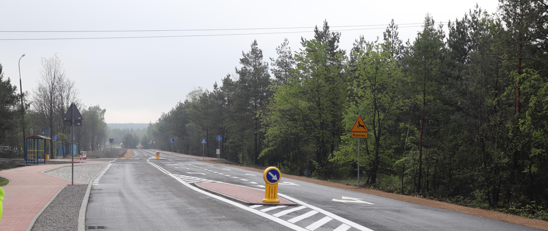Na zdjęciu widać nową drogę, po jednym pasie ruchu w każdą stronę. Jezdnie oddziela wysepka. Po prawej stronie widać wysokie, zielone drzewa. Po lewej stronie, w oddali widać zatokę i przystanek autobusowy. 