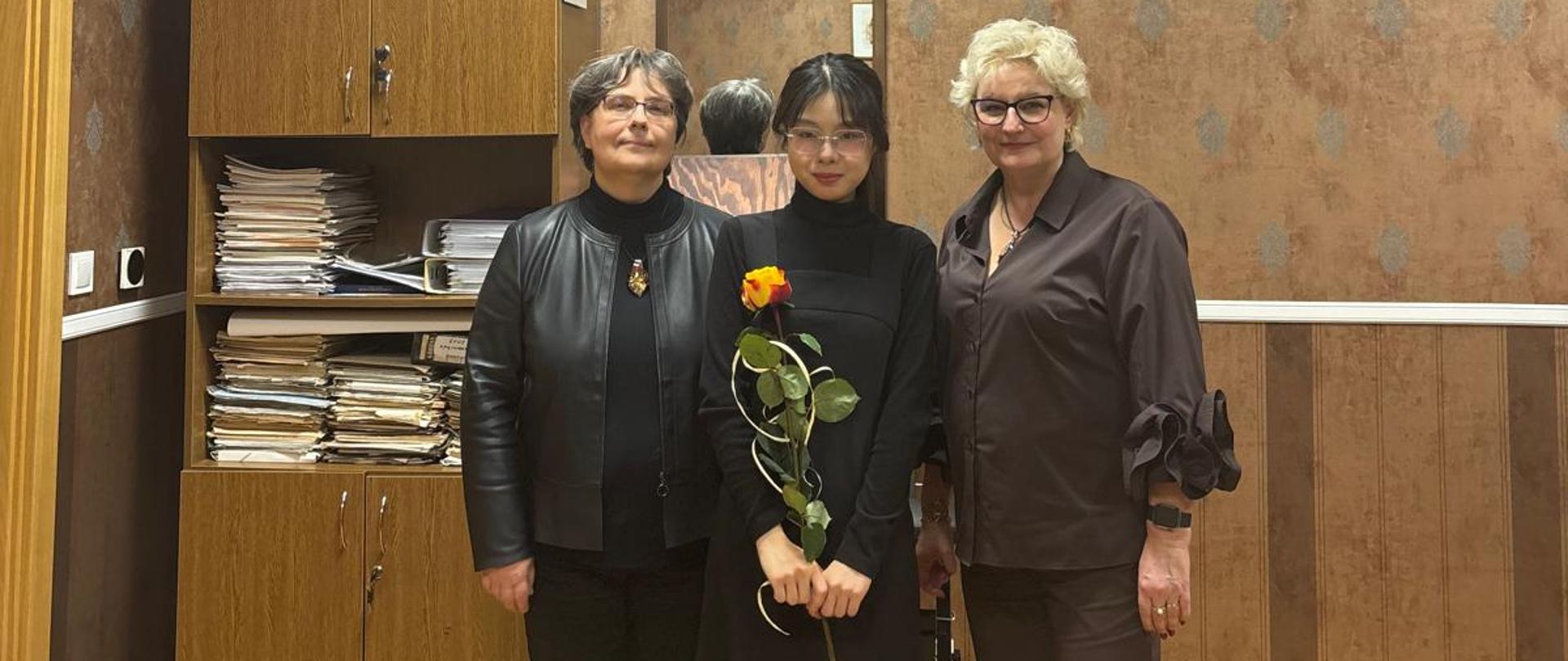 Na zdjęciu po prawej stronie kobieta blondynka ubrana na w brązowe spodnie i brązową koszulę. Obok niej młoda kobieta brunetka ubrana na czarno, która trzyma żółto- pomarańczową różę. Dalej do lewej strony kobieta w czarnych spodniach i czarnej marynarce. Tło obrazka w odcieniach ciemnobrązowych.