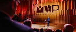 Mężczyzna w garniturze stoi za przezroczystą mównicą i mówi do mikrofonu, za nim oświetlona kolorowo ściana ze stylizowanymi literami MHP.