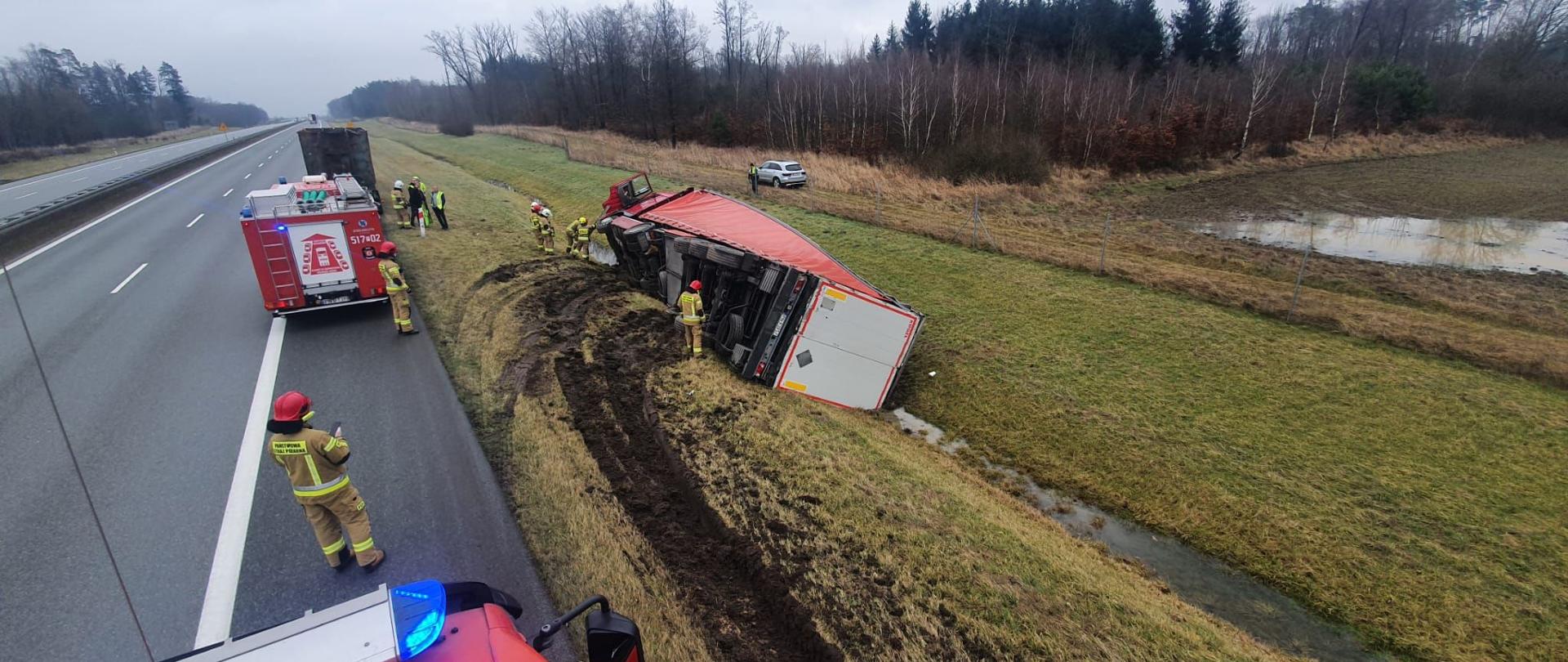 Dzień, TIR leżący na prawym boku poza jezdnią autostrady.