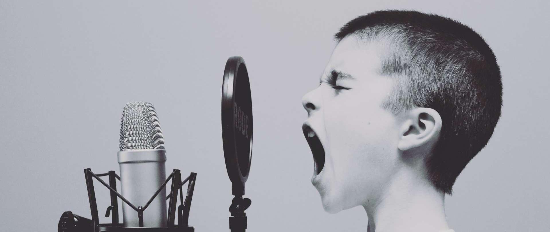 ciemnowłosy chłopiec ubrany w biały podkoszulek śpiewa do mikrofonu studyjnego