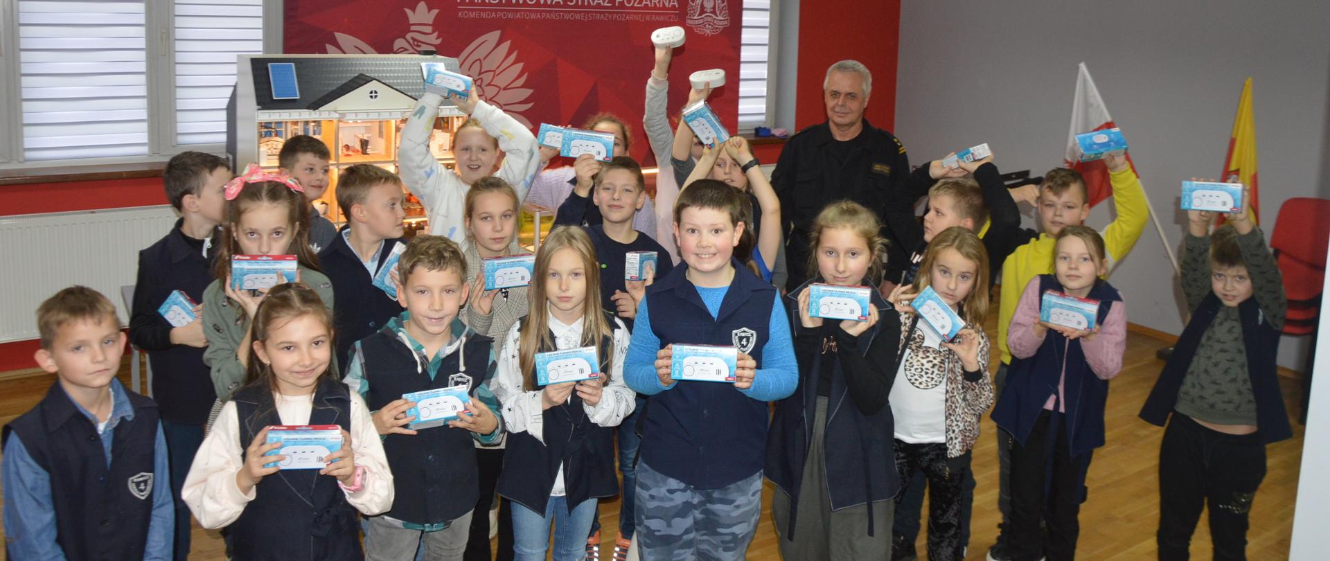 Trzecia edycja czadowego konkursu. Świetlica Komendy Powiatowej PSP w Rawiczu. Na tle symulatora zagrożeń pożarowych i ścianki reklamowej stoją uczniowie klasy IIID SP nr 4 w Rawiczu. i strażak. Dzieci trzymają w rękach czujniki tlenku węgla, Niektóre z nich unoszą urządzenia nad głową.