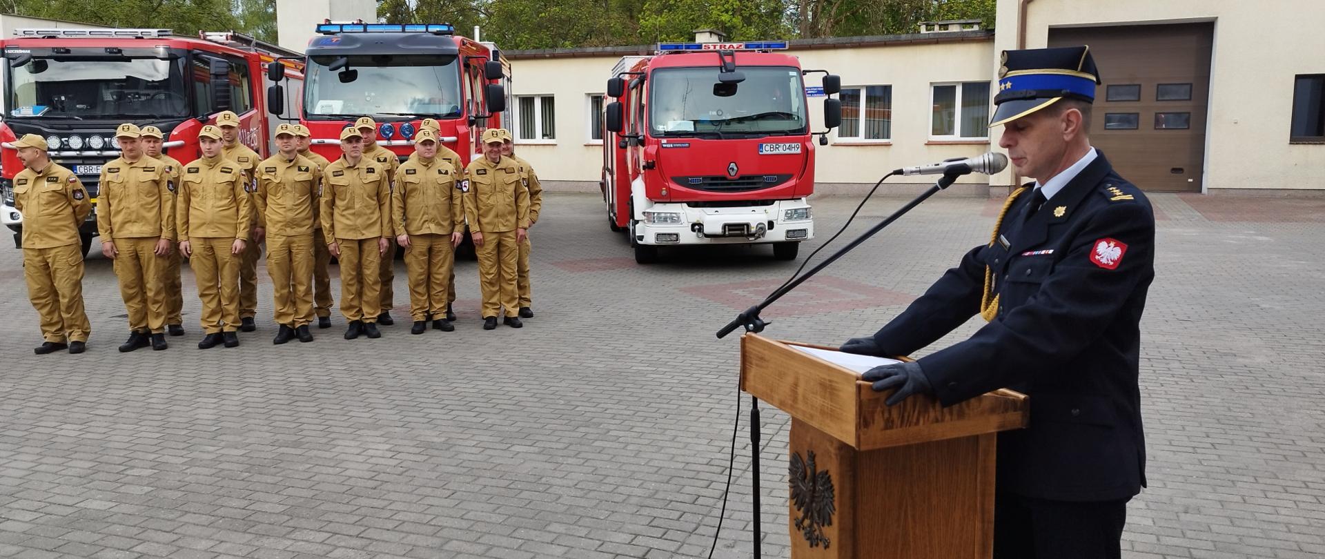 Zdjęcie przedstawia Komendanta Powiatowego PSP w Brodnicy st. bryg. Andrzeja Góreckiego przemawiającego przed uroczystym podniesieniem flagi państwowej na maszt. Na drugim planie strażacy Komendy w Brodnicy. Za nimi pojazdy gaśnicze.