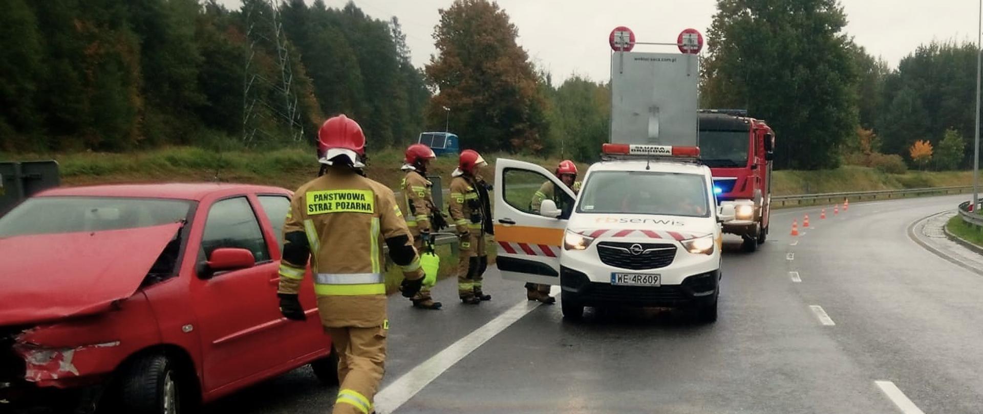 Zdjęcie przedstawia trasę S7, na której doszło do zderzenia samochodu z barierami. Z lewej strony, na poboczu stoi uszkodzone auto, na pasie drogowym zaparkował samochód przedstawiciela zarządcy drogi, za którym stoi samochód pożarniczy. 4 strażaków pracuje na terenie działań.