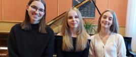 Zdjęcie kolorowe. Trzy uśmiechnięte dziewczyny siedzą przed otwartym fortepianem. Po lewej stronie siedzi dziewczyna w okularach, o ciemnych włosach, ubrana w czarny sweter. W środku siedzi dziewczyna o blond włosach, ubrana w czarny sweter. Po prawej stronie siedzi dziewczyna o blond włosach, ubrana w biały sweter.