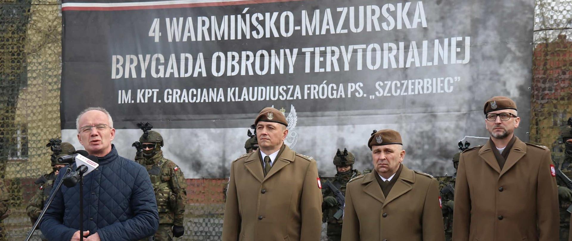 Na zdjęciu od prawej trzech w wojskowych galowych mundurach. Z lewej przemawia starszy pan w niebieskiej kurtce. W tle baner z napisem "4 warmińsko- mazurska brygada obrony terytorialnej".