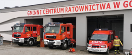 Trzy samochody strażackie wyjeżdżają z otwartych garaży budynku z napisem Gminne Centrum Ratownictwa
