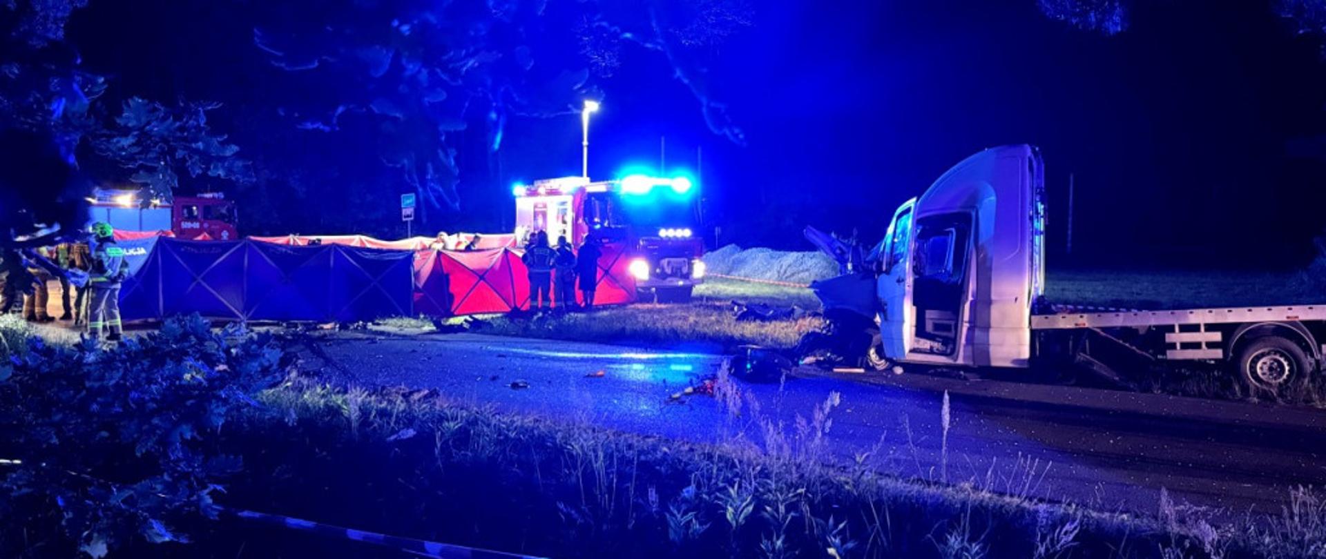 Nocna akcja ratunkowa straży pożarnej i policji przy oświetlonej drodze. Widoczna laweta oraz rozstawione parawany ochronne. Na miejscu pracują ratownicy w kaskach i samochody pożarnicze z włączonymi sygnałami świetlnymi.