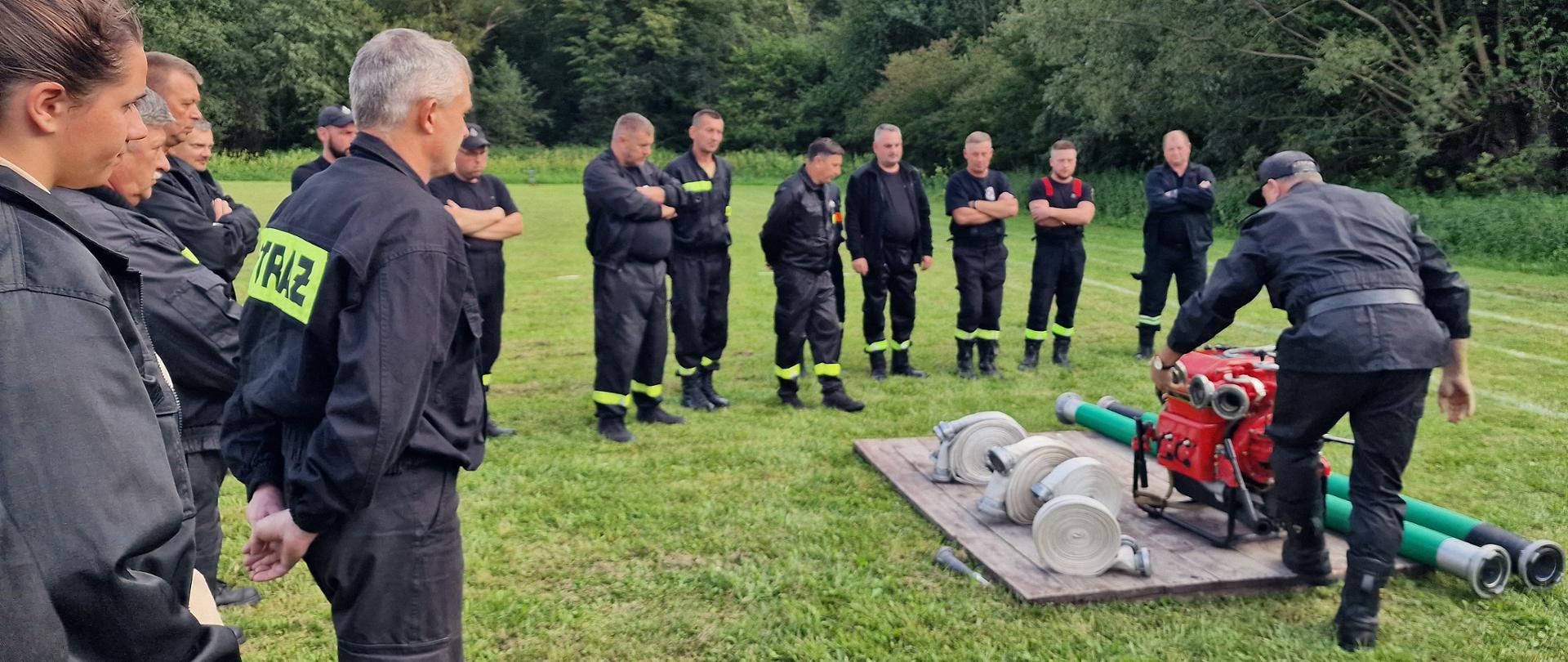 Widok z przodu. Prowadzący szkolenie (tyłem) stoi przy motopompie. Wokół niego zgromadzeni uczestnicy szkolenia (przodem).