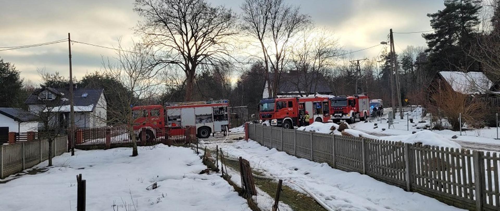 Zdjęcie obrazuje pojazdy Państwowej Straży Pożarnej z Komendy Powiatowej w Skarżysku-Kamiennej oraz pojazd z Ochotniczej Straży Pożarnej w Bliżynie, które stoją na ulicy przed palącym się budynkiem mieszkalnym jednorodzinnym.