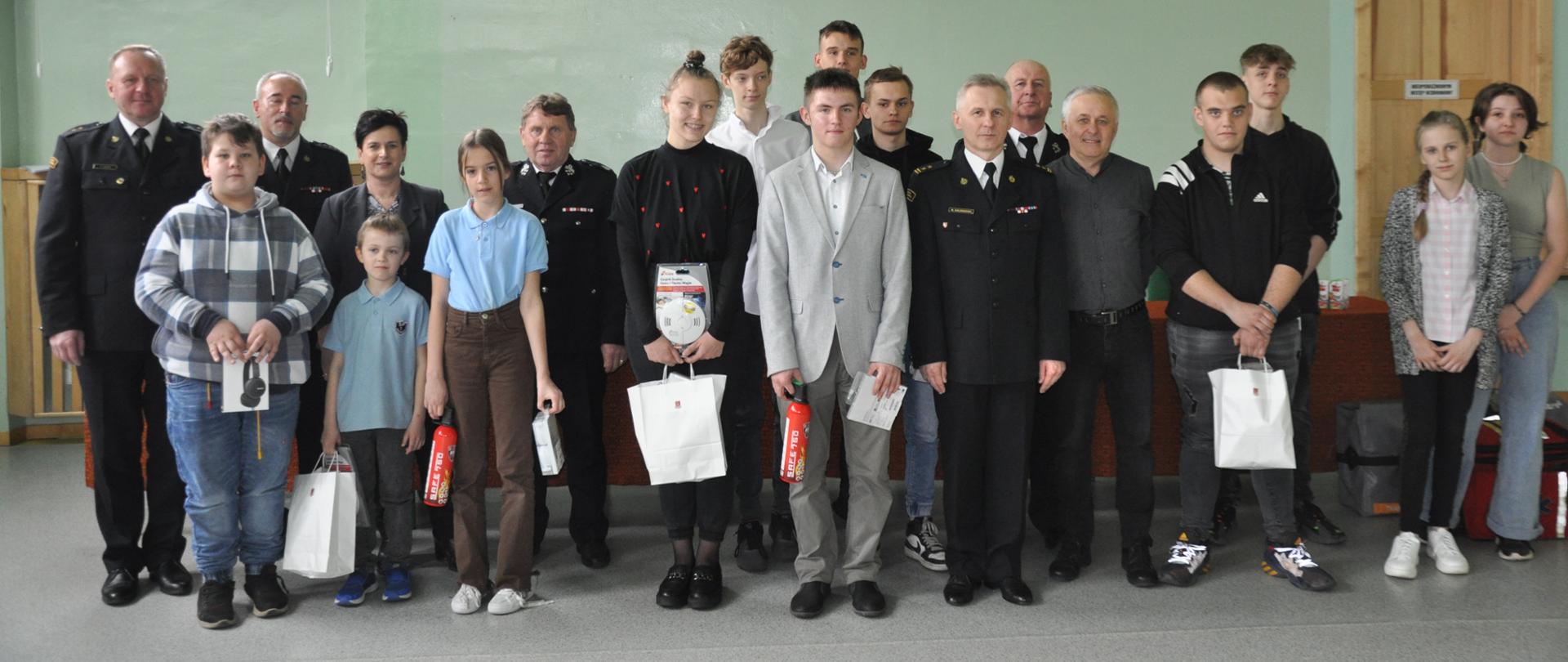 Uczestnicy - uczniowie oraz komisja - złożona z funkcjonariuszy PSP i OSP, zdjęcie grupowe. Na pierwszym tle uczestnicy ze zdobytymi nagrodami. 