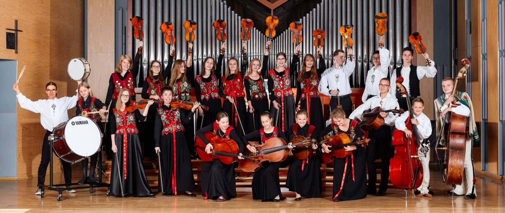 Zdjęcie przestawia skład orkiestry szkolnej. W dwóch rzędach stoją z przodu od lewej do prawej bębniarze, dwóch skrzypków wiolonczeliści, kontrabasiści. W drugim rzędzie skrzypkowi. Wszyscy podnoszą instrumenty do góry robiąc przy tym zabawne miny.