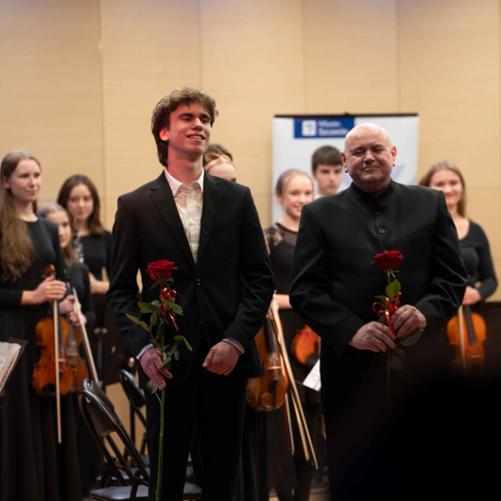 zdjęcie przedstawia ucznia szkoły oraz dyrygenta orkiestry trzymających kwiaty róży w dłoniach podczas oklasków po wykonaniu utworu, w tle orkiestra szkoły w pozycji stojącej oraz baner miasta Szczecin