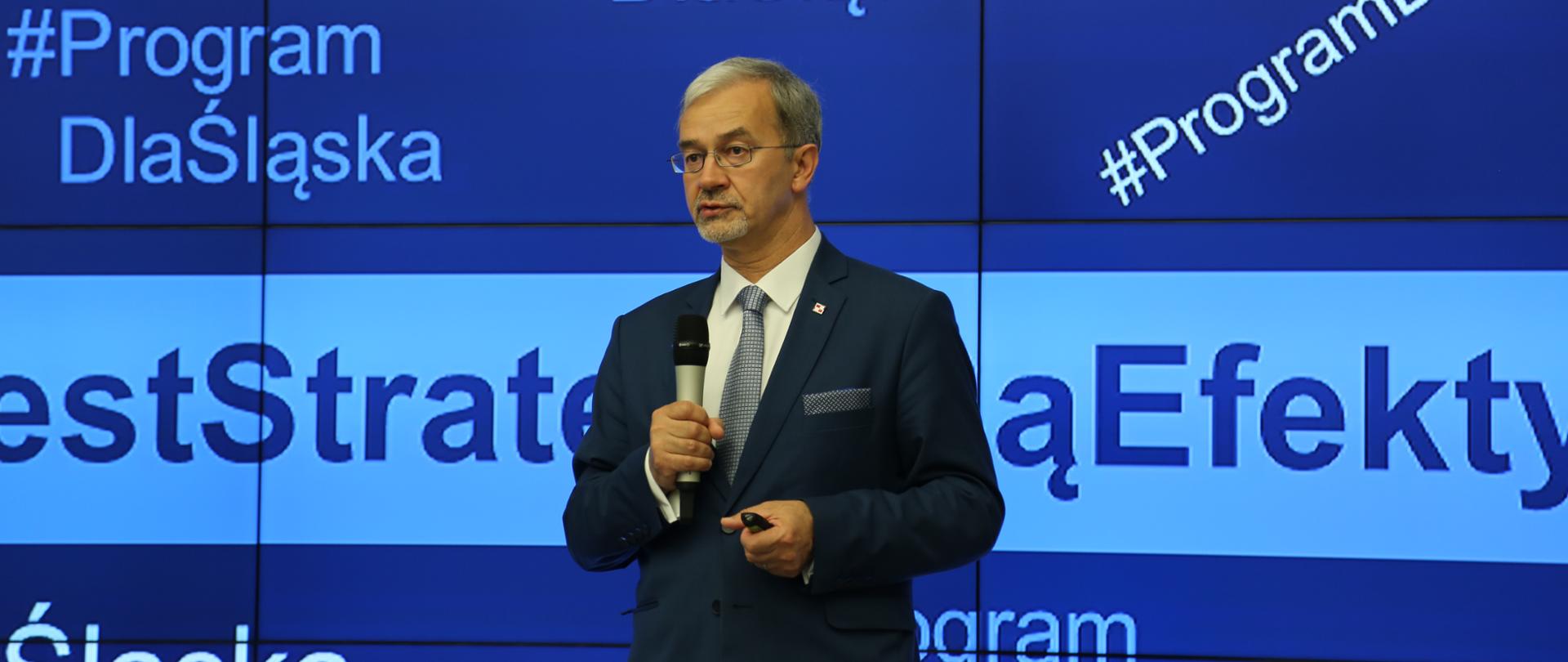 Na tle ekranu z niebieskim tłem i napisami Program dla Śląska stoi minister Jerzy Kwieciński i mówi do mikrofonu, który trzyma w ręku