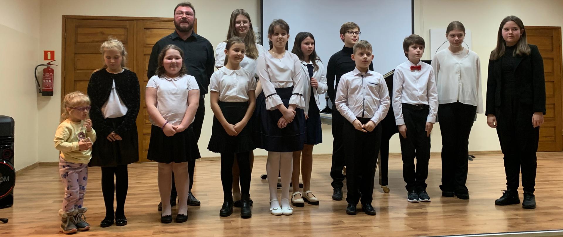 Na scenie wykonawcy koncertu z klasy fortepianu p. Piotra Wilka wraz z nauczycielem. Wszyscy stoją w dwóch rzędach ubrani na galowo. W tle fragmenty czarnego fortepianu. 