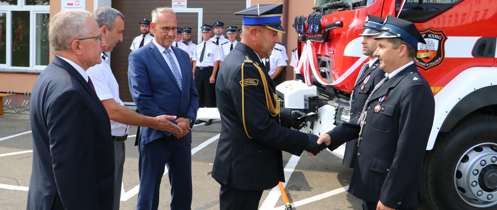 Kolorowa fotografia wykonana na zewnątrz w pogodny dzień. Przedstawia strażaka ubranego w mundurowaniu galowym PSP wręczający odznaczenia strażakom OSP wraz osobami cywilnymi. W tle nowy samochód pożarniczy przygotowany do poświęcenia.