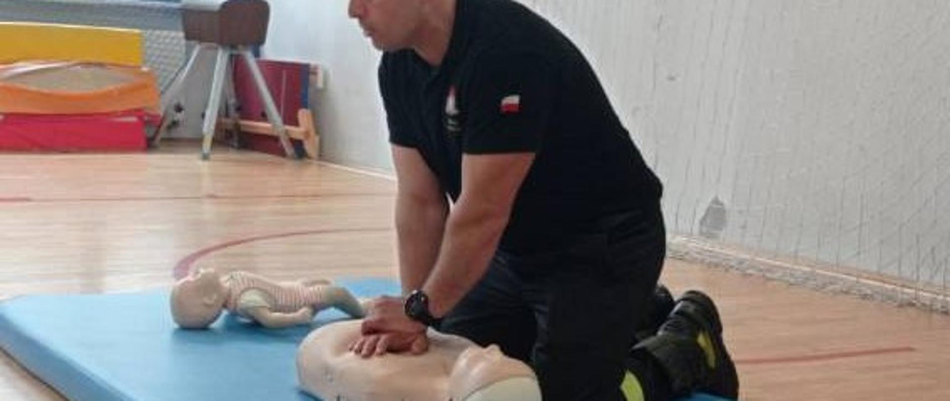 Na zdjęciu przedstawiono warsztaty z uczniami w szkole podstawowej w ramach profilaktyki bezpieczeństwa. Ratownik wykonuje masaż serca na fantomie ćwiczebnym.