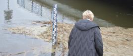 Okolice miejscowości Słupia Kapitulna. Na wale przeciwpowodziowym rzeki Orla stoi kobieta, przedstawicielka gminnego centrum zarzadzania kryzysowego, która z wodowskazu dokonuje odczytu poziomu wody w rzece. Z prawej strony widoczny jest fragment mostu. Na powierzchni lustra wody znajdują się zanieczyszczenia w postaci roślinności.