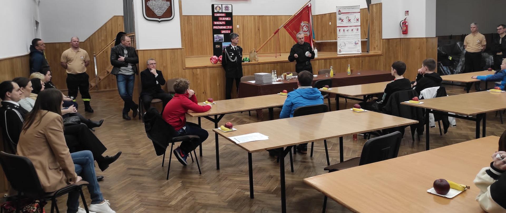 Kolorowa fotografia wykonana wewnątrz pomieszczenia. Przedstawia ona dużą jasną salę, na której stoją krzesła i stoliki. Przy nich siedzi kilkanaście młodych osób. Na końcu sali stół i dwóch umundurowanych strażaków. Jeden z nich trzyma biały przedmiot i mówi do zebranych. Po lewej stronie pod ścianą siedzi kilka cywilnych osób a po prawej stoją umundurowani strażacy. W tle scena ze sztandarem i banerami informacyjnymi.