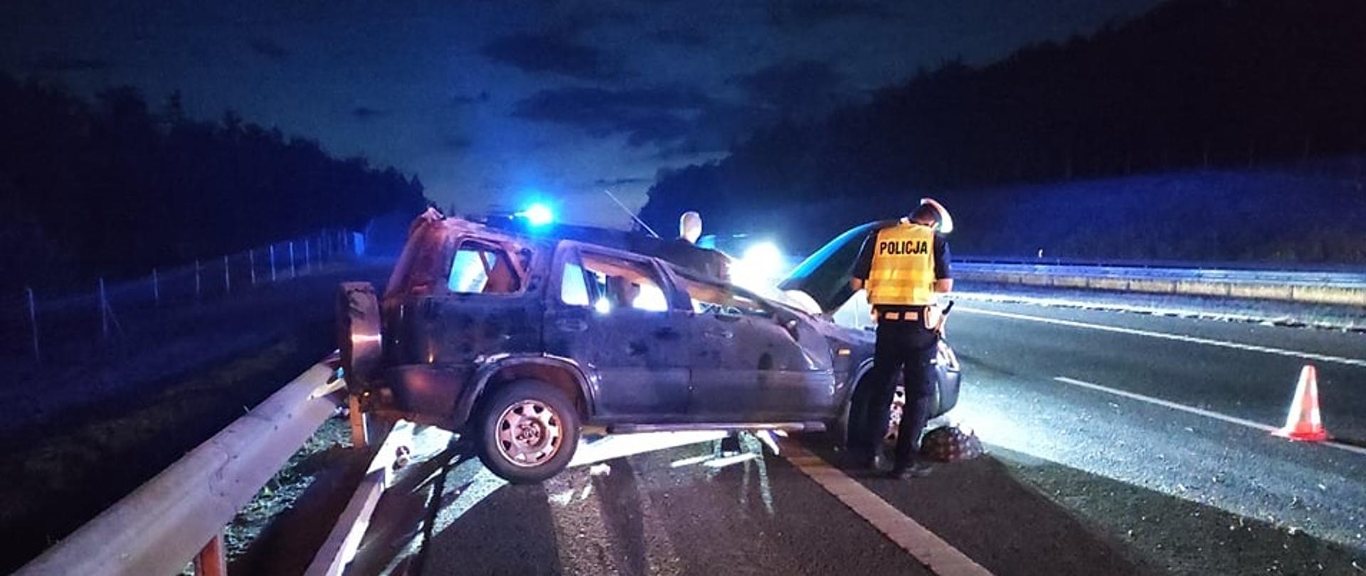 Na zdjęciu widać uszkodzony pojazdu marki Honda po zderzeniu z barierą energochłonną. Przy pojeździe stoi funkcjonariusz Policji. Za pojazdem widać radiowóz policyjny, oraz samochód pożarniczy, które mają włączone światła błyskowe.