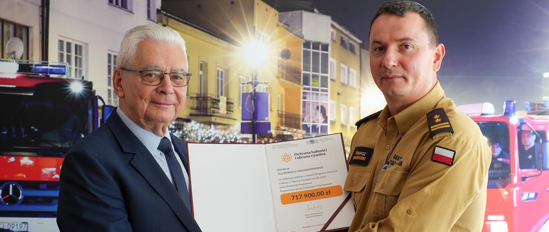 Starosta Ciechanowski i Komendant Powiatowy podający sobie prawą dłoń. Komendant w stroju służbowym trzyma przekazaną promesę na dotację dla jednostki ze środków programu ochrony ludności i obrony cywilnej 