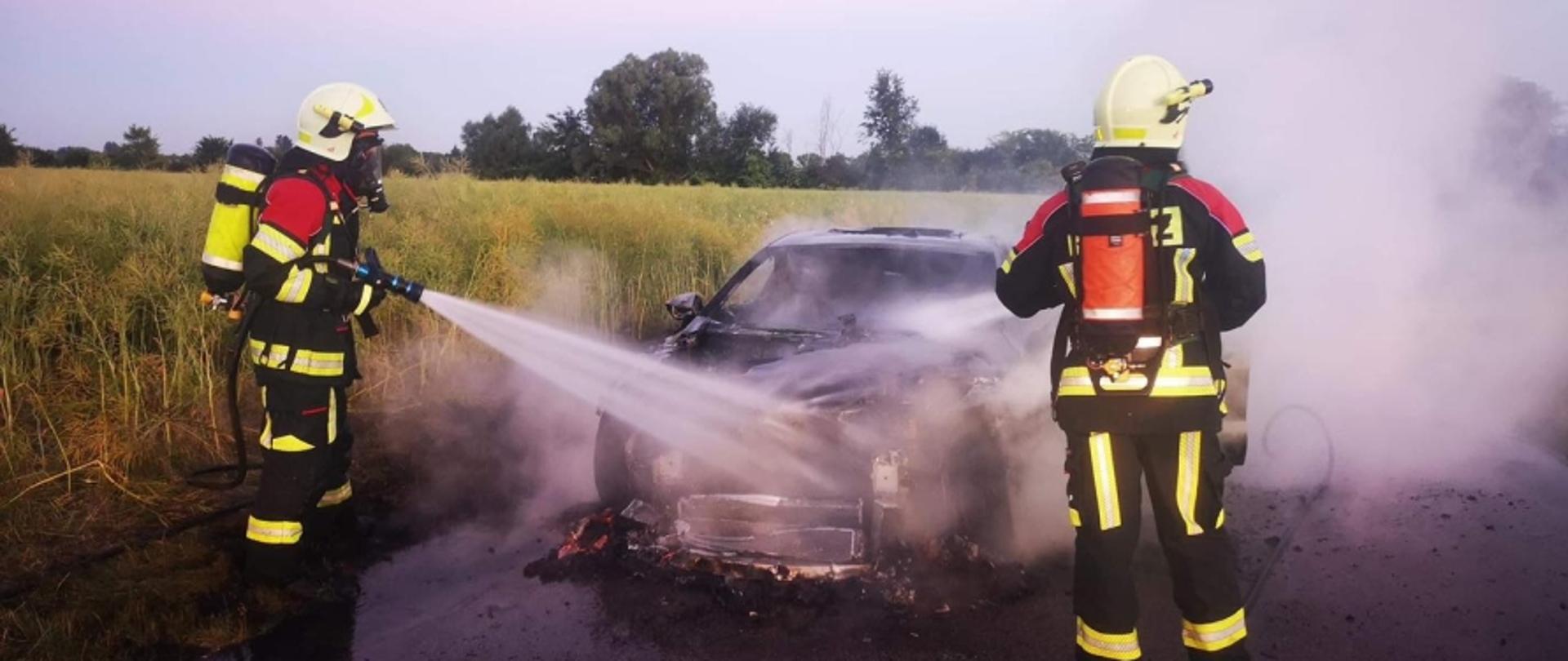 Na zdjęciu widoczny spalony samochód osobowy stojący na jezdni, który dogasza dwóch strażaków w czerwono-czarnych ubraniach specjalnych, białych hełmach, maskach na twarzy i butlach na plecach.