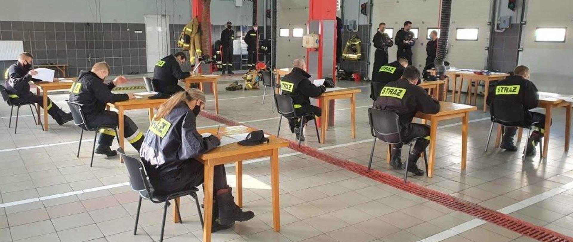 Na zdjęciu widać trwającą część teoretyczną egzaminu strażaków ratowników OSP. Egzaminowanych jest siedmiu, podczas gdy pilnujących jest pięciu. Wszyscy obecni na sali egzaminacyjnej, za którą służy garaż, ze względu na panujący stan pandemii i wymagania reżimu sanitarnego mają na twarzach maseczki. Garaż ma podłogę wyłożoną szarymi płytkami przedzielonymi co pewien dystans płytkami czerwonymi a następnie białymi. Przez środek pomieszczenia widać czerwoną kratkę odwodnienia. Na ścianie widoczne są zamontowane wrota garażowe koloru białego, wrota posiadają na trzecim przęśle cztery okna. W tle zdjęcia widać dwa filary podtrzymujące strop, do wysokości 1,5 metra filary obłożone są szarymi płytkami, a do sufitu pomalowane są na czerwono. Na ścianie widocznej w tle znajdują się białe drzwi wejściowe, ściana do wysokości drzwi pokryta jest ciemnoszarymi płytkami, podczas gdy wyżej pomalowana jest na kolor jasnoszary. Egzaminowani siedzą na czarnych krzesłach przy drewnianych jasnobrązowych pojedynczych stolikach, które oddalone są od siebie o 2 metry. Wszyscy obecni na sali ubrani są w czarne ubrania koszarowe z napisem straż na plecach.
