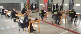Na zdjęciu widać trwającą część teoretyczną egzaminu strażaków ratowników OSP. Egzaminowanych jest siedmiu, podczas gdy pilnujących jest pięciu. Wszyscy obecni na sali egzaminacyjnej, za którą służy garaż, ze względu na panujący stan pandemii i wymagania reżimu sanitarnego mają na twarzach maseczki. Garaż ma podłogę wyłożoną szarymi płytkami przedzielonymi co pewien dystans płytkami czerwonymi a następnie białymi. Przez środek pomieszczenia widać czerwoną kratkę odwodnienia. Na ścianie widoczne są zamontowane wrota garażowe koloru białego, wrota posiadają na trzecim przęśle cztery okna. W tle zdjęcia widać dwa filary podtrzymujące strop, do wysokości 1,5 metra filary obłożone są szarymi płytkami, a do sufitu pomalowane są na czerwono. Na ścianie widocznej w tle znajdują się białe drzwi wejściowe, ściana do wysokości drzwi pokryta jest ciemnoszarymi płytkami, podczas gdy wyżej pomalowana jest na kolor jasnoszary. Egzaminowani siedzą na czarnych krzesłach przy drewnianych jasnobrązowych pojedynczych stolikach, które oddalone są od siebie o 2 metry. Wszyscy obecni na sali ubrani są w czarne ubrania koszarowe z napisem straż na plecach.