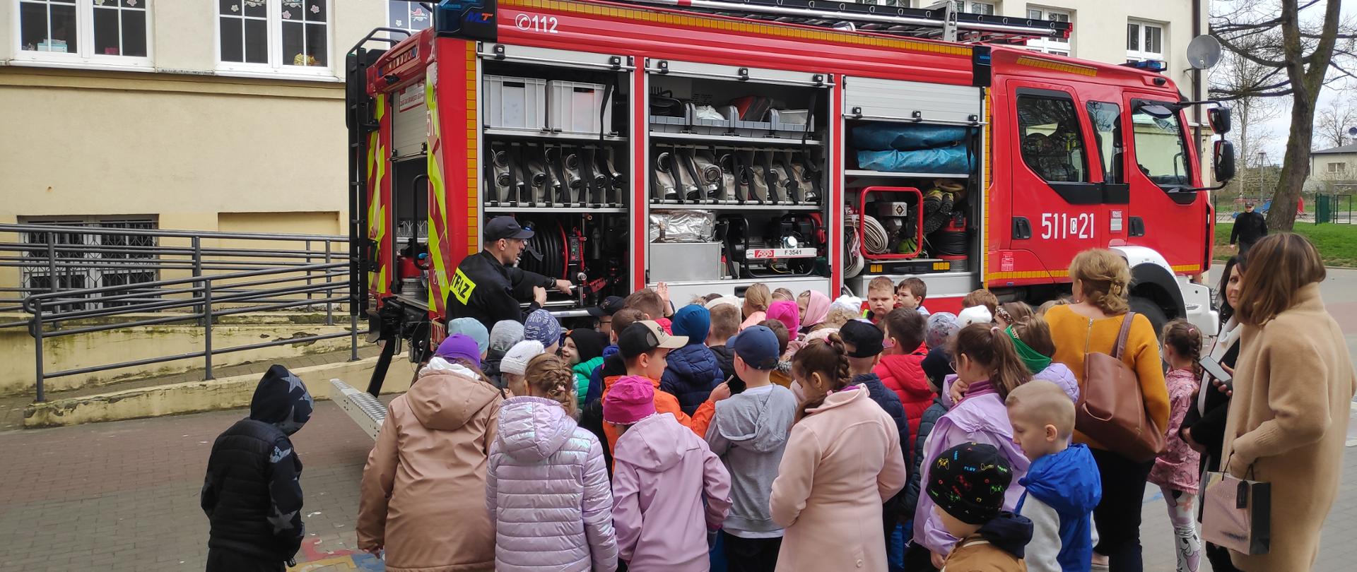 Grupa dzieci wraz z opiekunami znajduje się na placu Szkoły Podstawowej numer jeden w Rypinie. Funkcjonariusz Państwowej Straży Pożarnej przedstawia im samochód pożarniczy wraz z jego wyposażeniem. W tle budynek szkoły oraz plac zabaw.
