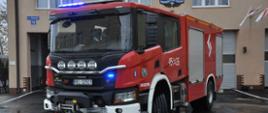 Na wyasfaltowanym placu stoi samochód strażacki z włączonymi światłami barwy niebieskiej. W tle część budynku z oknami i bramami garażowymi.