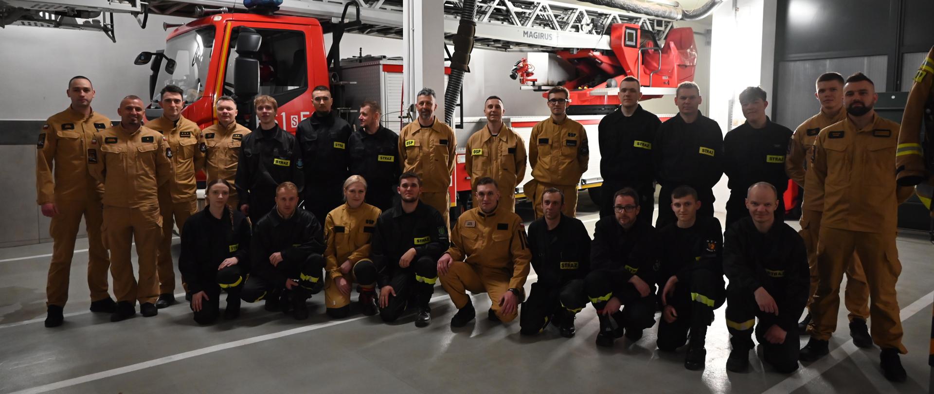 Grupa strażaków w ubraniach służbowych pozuje do wspólnego zdjęcia w garażu remizy, na tle wozu strażackiego z drabiną mechaniczną.