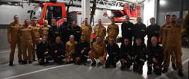 Grupa strażaków w ubraniach służbowych pozuje do wspólnego zdjęcia w garażu remizy, na tle wozu strażackiego z drabiną mechaniczną.