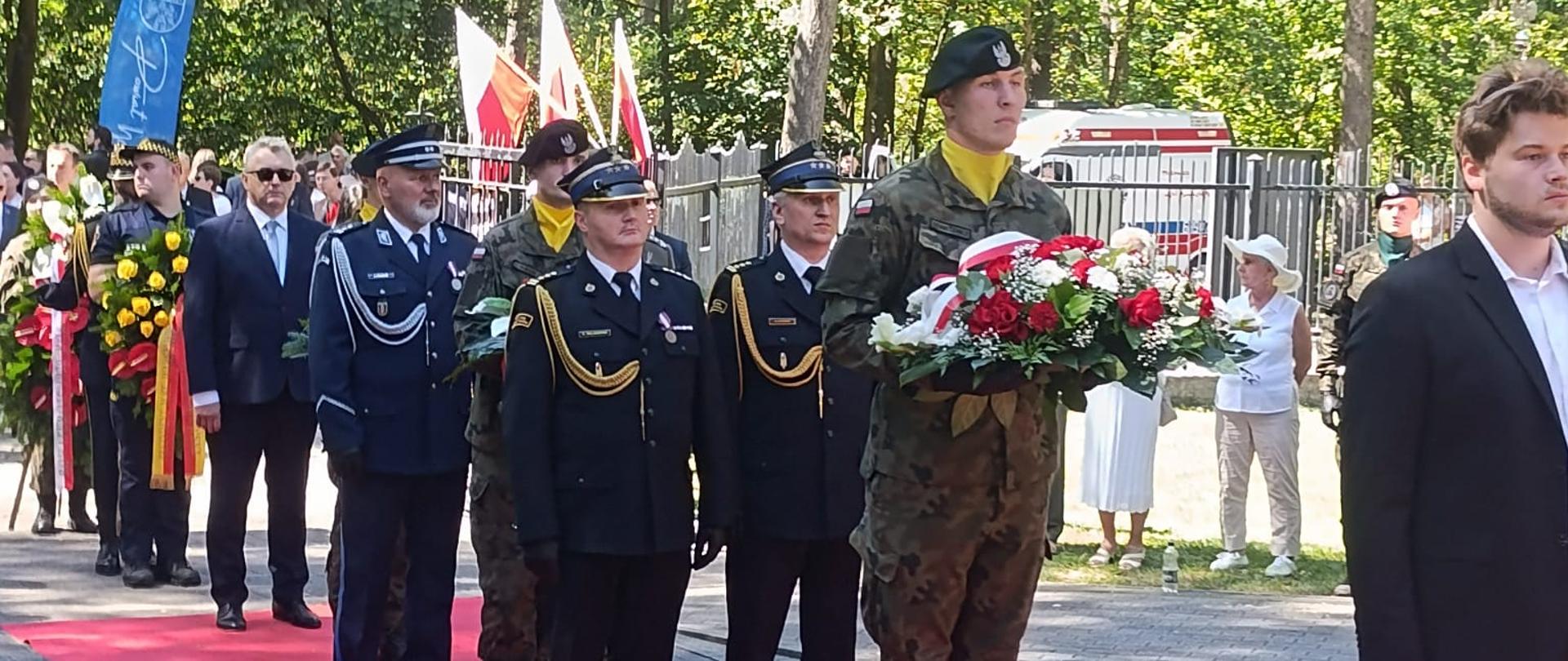 Przedstawiciele służb mundurowych oraz delegacje z wieńcami podczas uroczystości rocznicowych na terenie leśnym.