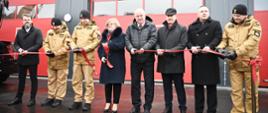 Uczestnicy uroczystości stoją przed strażnicą PSP po przecięciu czerwonej wstęgi. W tle widoczne czerwone bramy garażowe budynku.