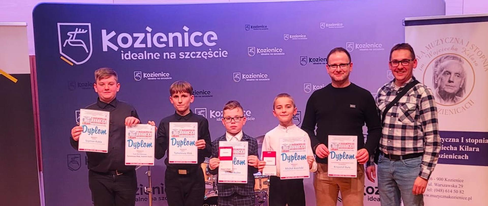 Zdjęcie laureatów stojących na scenie obok swoich nauczycieli. Uczniowie trzymają przed sobą zdobyte dyplomy, a za nimi widoczny jest baner wydarzenia oraz rollup promujący miasto Kozienice