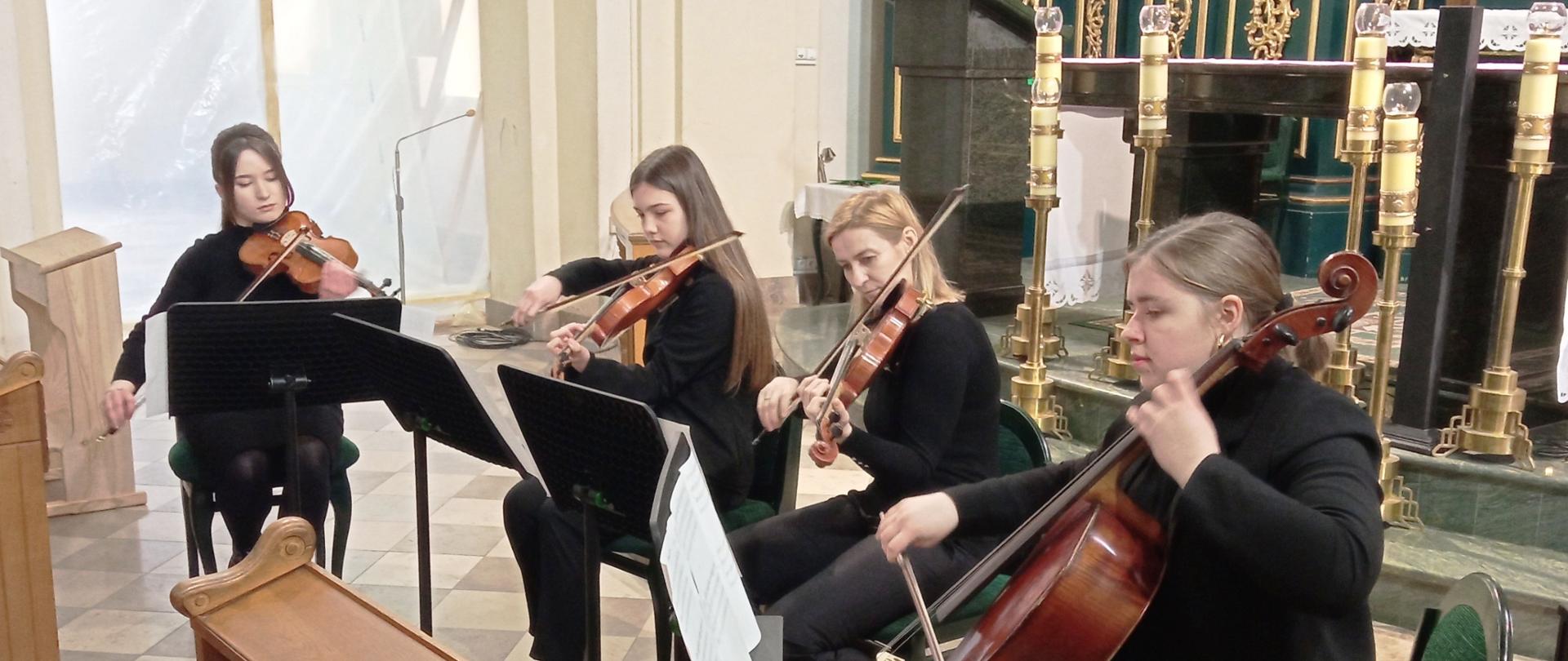 Zdjęcie przedstawiające siedzące na krzesłach przy pulpitach, występujące przed ołtarzem Trio smyczkowe