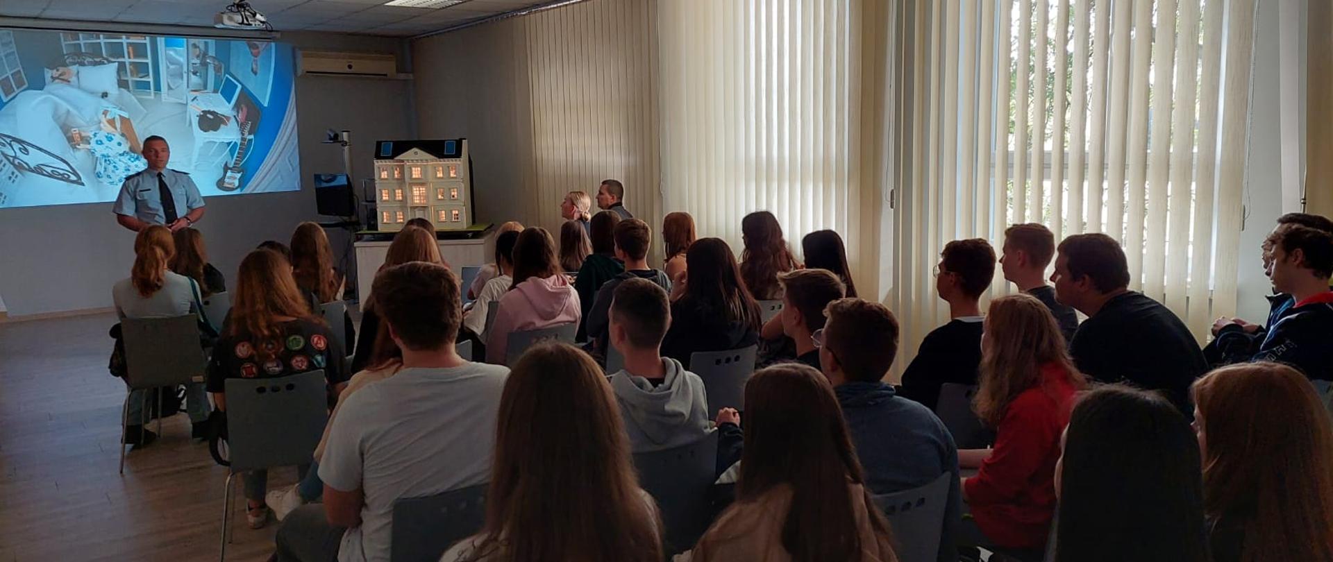 Zdjęcie przedstawia strażaka, który na sali komendy przedstawia młodzieży z klas ratowniczych zagrożenia występujące w prywatnych budynkach mieszkalnych przy wykorzystaniu mobilnego symulatora zagrożeń pożarowych. Młodzież siedzi na krzesłach. Strażak stoi z tyłu na ścianie wyświetlane są pomieszczenia domku. 