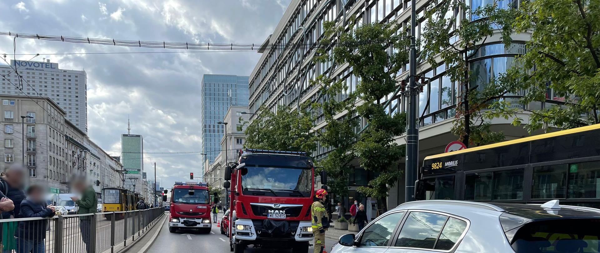 Na zdjęciu widać biały samochód marki Kia Sportage, który brał udział w zdarzeniu . Na dalszym planie widać dwa samochody ratowniczo-gaśnicze w kolorze czerwonym z elementami białego oraz ratownika w piaskowym ubraniu specjalnym zabezpieczający miejsce zdarzenia. 