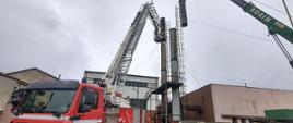 Na zdjęciu widać rozstawiony podnośnik straży pożarnej obok stalowego komina i dźwig unoszący przy pomocy zawiesi jego odciętą górną część 