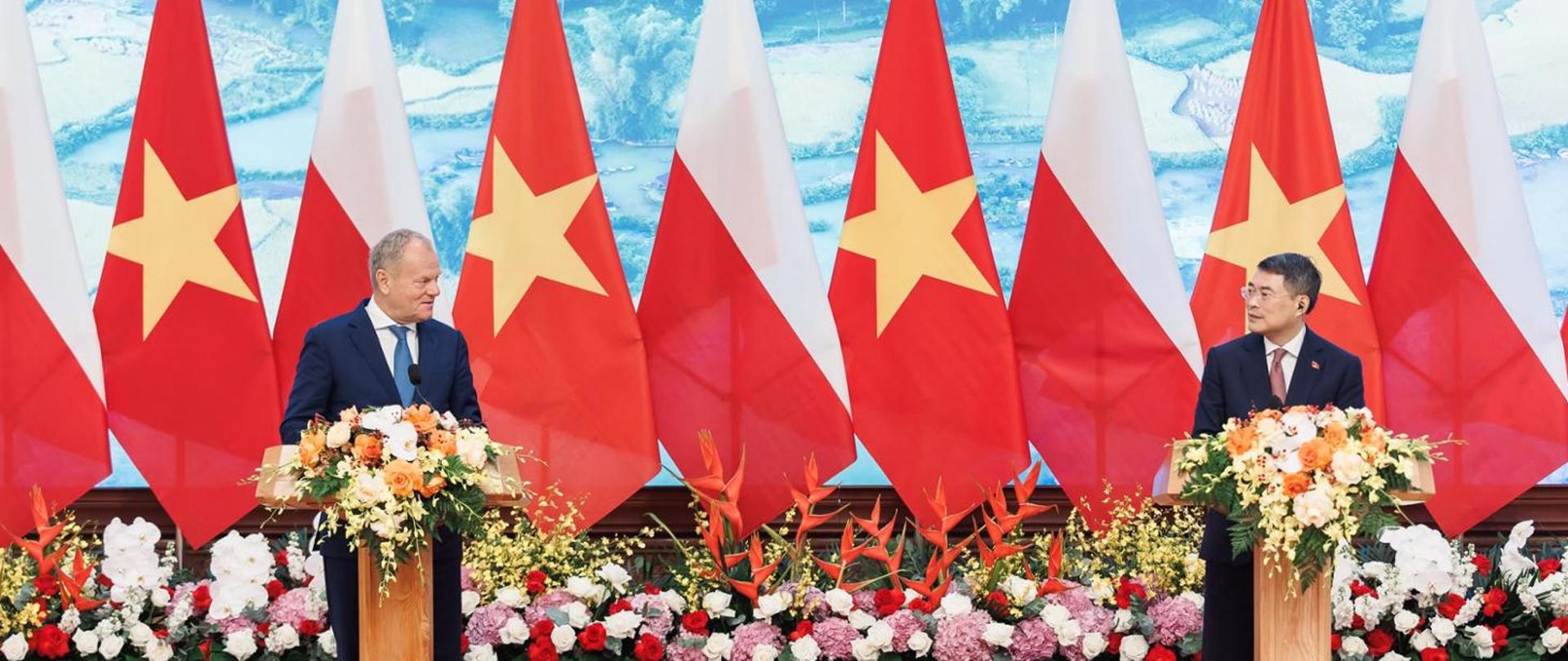 Premier D. Tusk (z lewej) i premier Lê Minh Hưng (z prawej) za mównicami podczas konferencji prasowej. Z przodu i na mównicach obfita dekoracja kwiatowa, w tle flagi Polski i Wietnamu