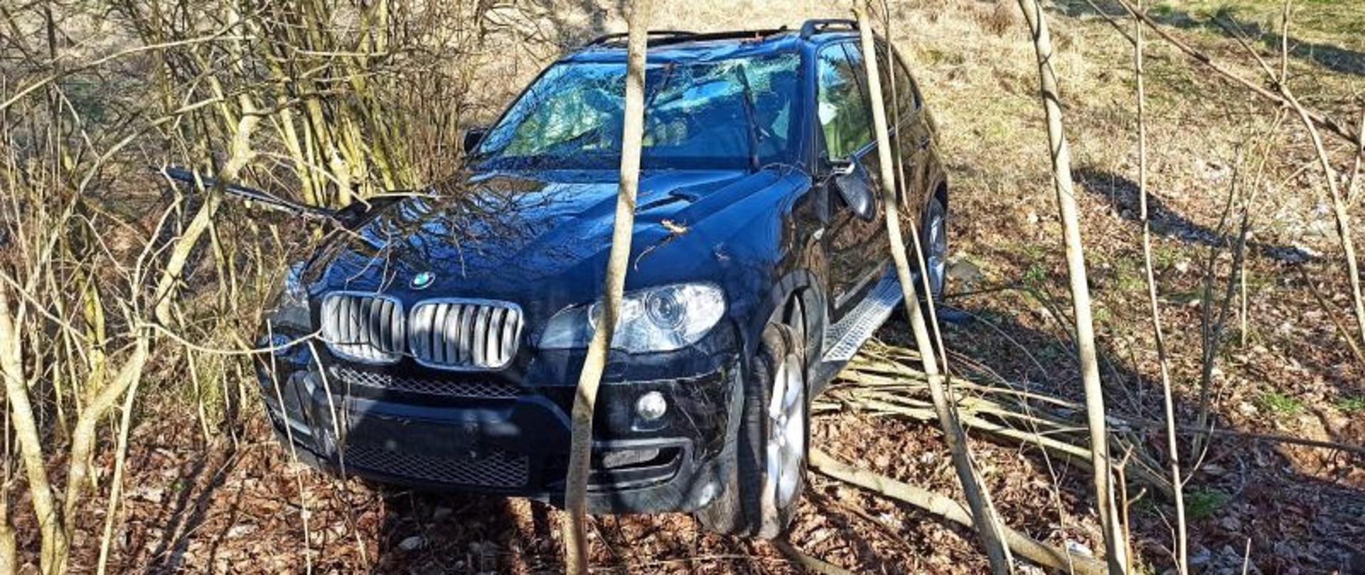 Zdjęcie przedstawia auto marki BMW. Jest to duży ciemny SUV. Auto stoi poza drogą, na kołach. Ma uszkodzony dach i wystrzelone poduszki powietrzne.