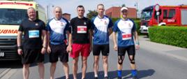 Czterech ubranych na sportowo mężczyzn stoi obok siebie na drodze asfaltowej, w tle widoczny żółty ambulans straży pożarnej i samochód gaśniczy