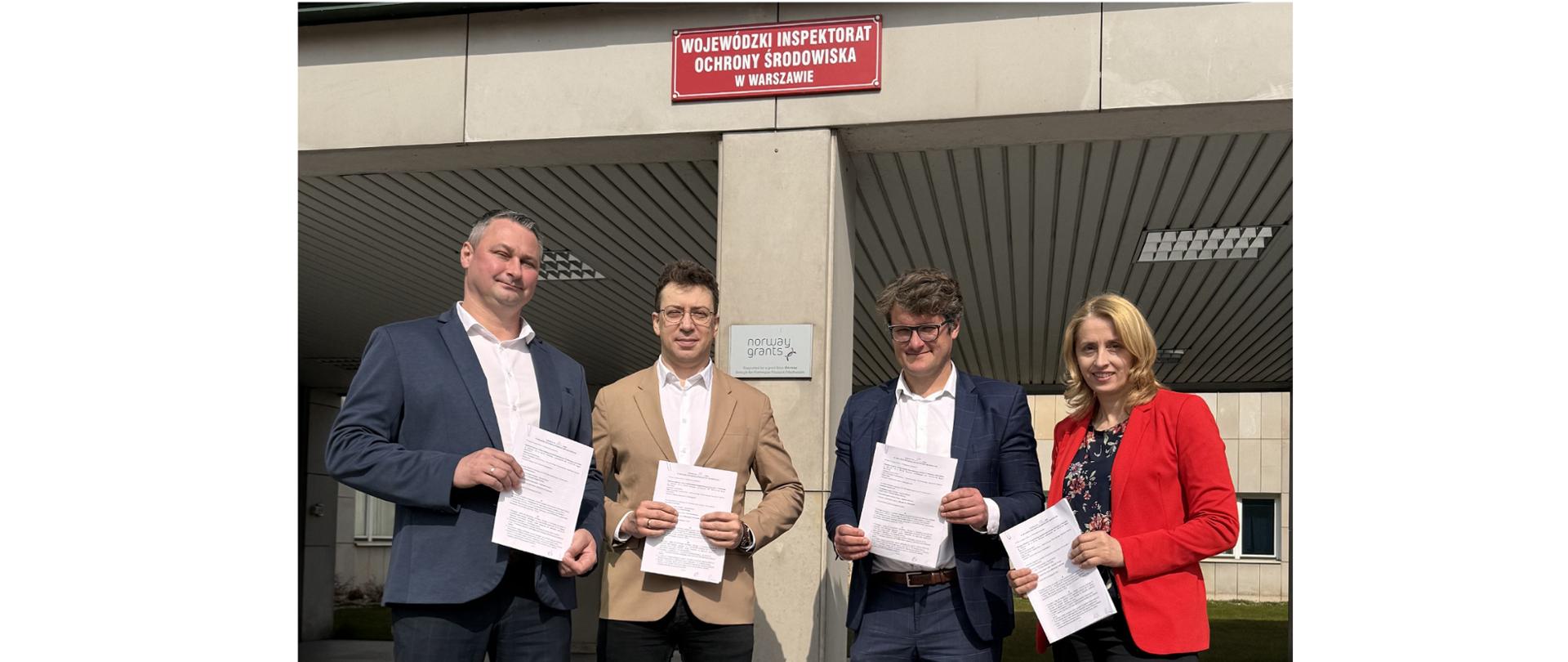 Cztery osoby pozują do pamiątkowego zdjęcia na tle budynku, na którym zawieszona jest tabliczka z napisem ”Wojewódzki Inspektorat Ochrony Środowiska w Warszawie”. Osoby te trzymają w rękach podpisaną umowę użyczenia przestrzeni biurowej.
Pozują od lewej:
p. Krzysztof Cząstkiewicz, Mazowiecki Wojewódzki Inspektor Ochrony Środowiska,
p. Paweł Łukaszuk, p.o. Kierownika Delegatury WIOŚ w Warszawie w Mińsku Mazowieckim,
p. Remigiusz Lesław Górniak, Starosta Miński,
p. Marta Wojtaczka, I Zastępca Mazowieckiego Wojewódzkiego Inspektora Ochrony Środowiska.
