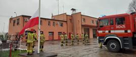 Na zdjęciu widać poczet flagowy. Ubrany jest w ubrania specjalne koloru piaskowego oraz czerwone hełmy. Trzech strażaków stoi przy maszcie i wciąga biało-czerwoną flagę na maszt. W tle widać budynek Jednostki Ratowniczo-Gaśniczej nr 2 koloru łososiowego. Budynek jest dwukondygnacyjny, pierwsza kondygnacja to czerwone i po części przeszklone bramy wyjazdowe. Druga kondygnacja to linia okien. z prawej strony kadru widzimy przednią część wozu strażackiego koloru czerwonego. Na tylnych drzwiach numery operacyjne w kolorze białym: trzysta czterdzieści dwa p dwadzieścia pięć". Strażacy stoją na placu, który jest wyłożony płytami betonowymi koloru szarego.
