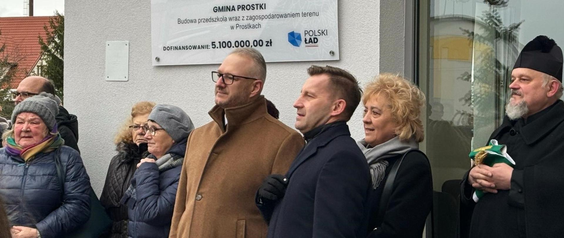 Grupa ludzi na tle nowo-wybudowanego przedszkola, obserwują moment przecięcia wstęgi